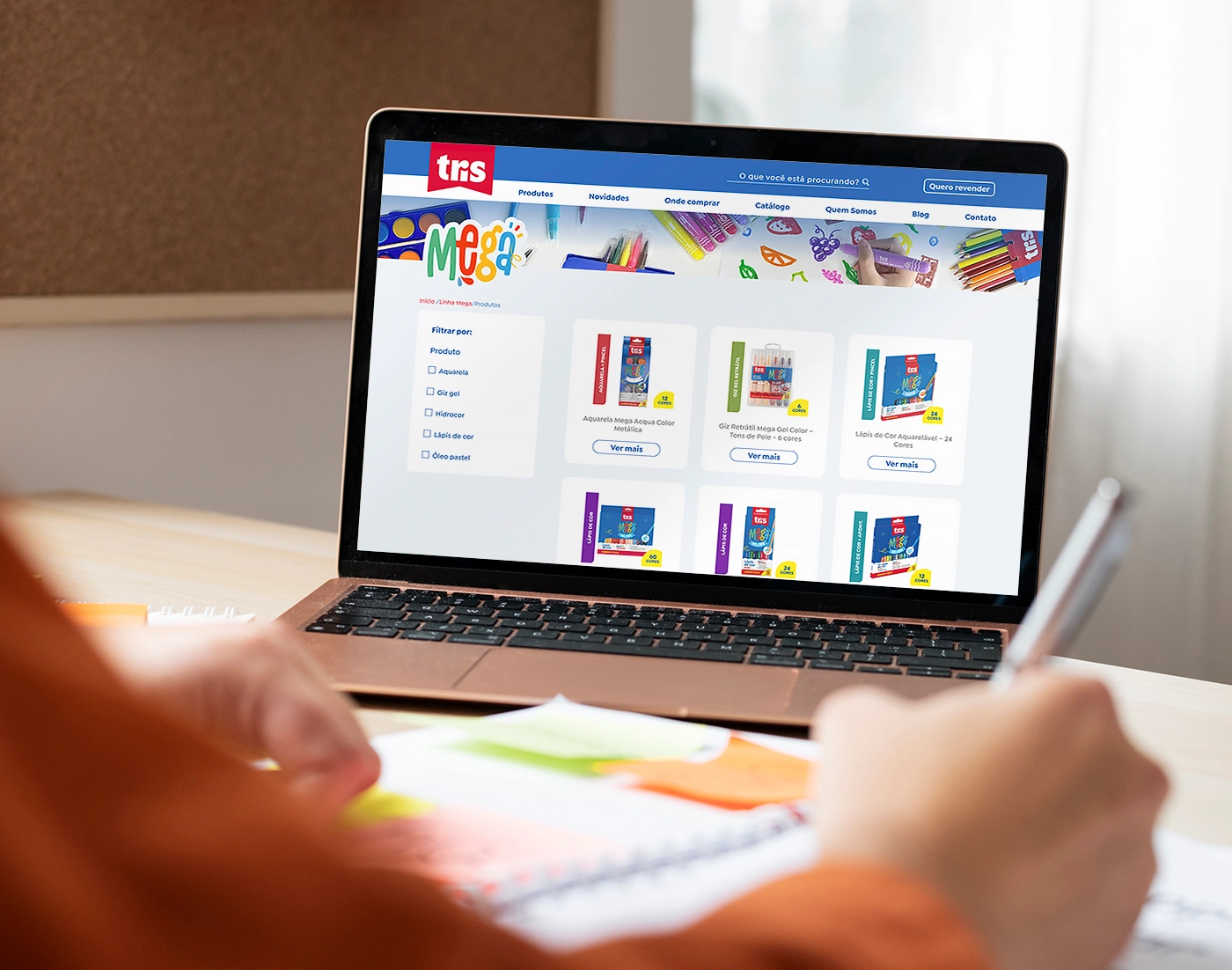 A laptop displaying the 'Tris Mega' website, showcasing colorful stationery items organized in a grid. The site features a variety of school supplies, including colored pencils and markers. In the background, a workspace with notebooks and a pen is visible.