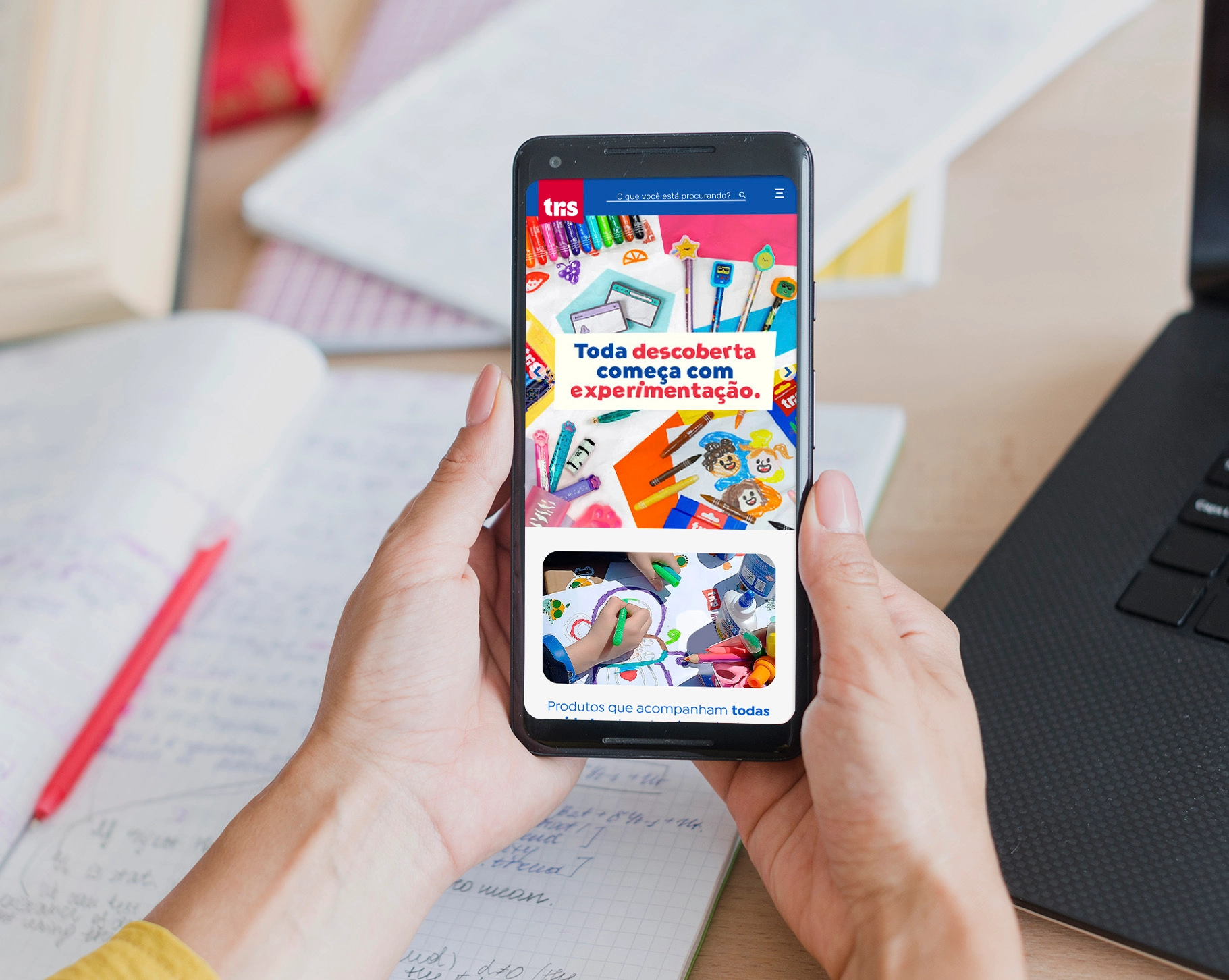 Person holding a smartphone displaying the homepage of the Tris website, which uses a primary color palette dominated by red and blue. The main banner features a bright, colorful photo montage of crayons, markers, and other art supplies surrounding the central text: 'Every discovery begins with experimentation'.