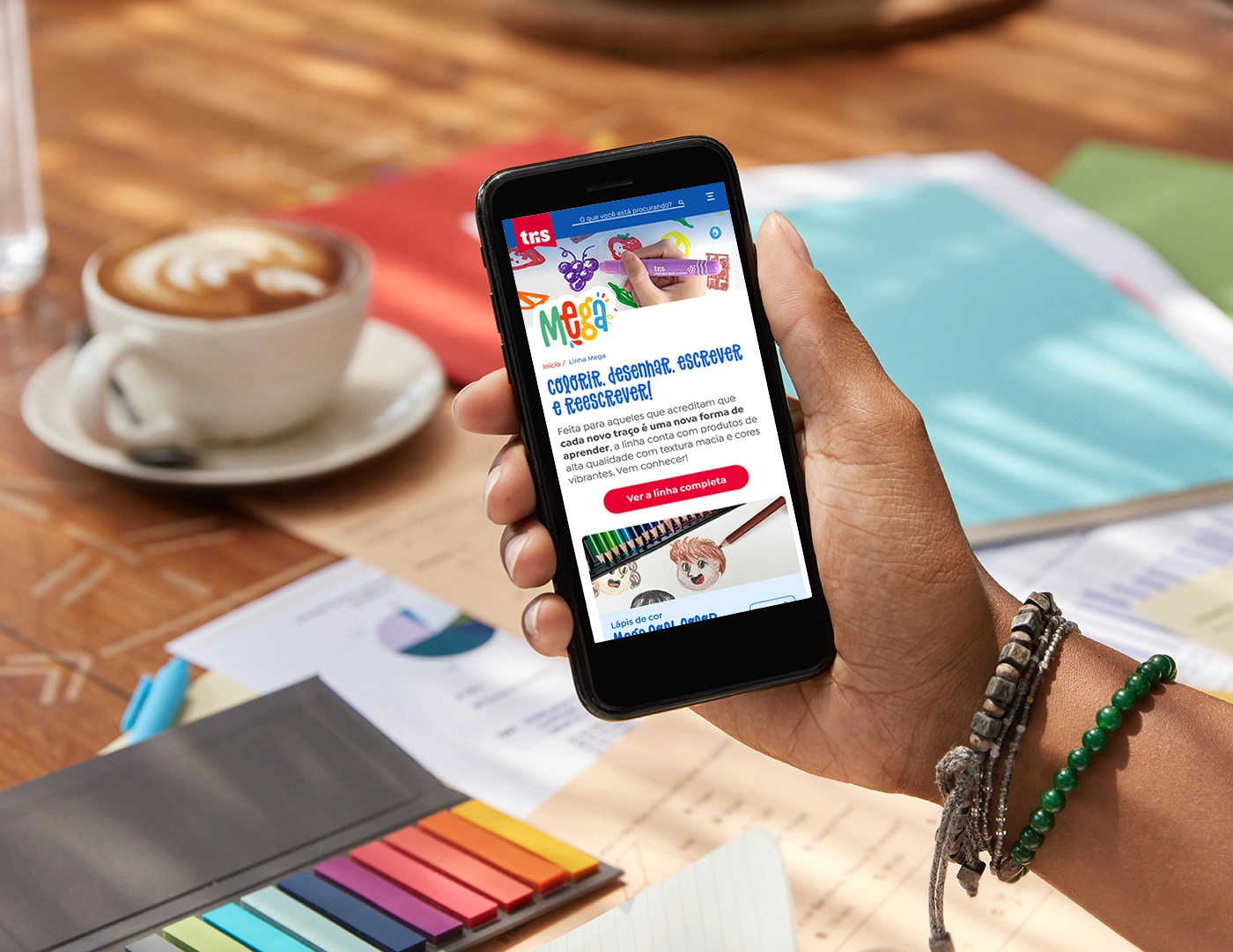 Person holding a smartphone displaying the 'Tris Mega' sub-brand page, with its headline 'Color, Draw, Write, and Rewrite!', a description of its purpose and a button to access all its products