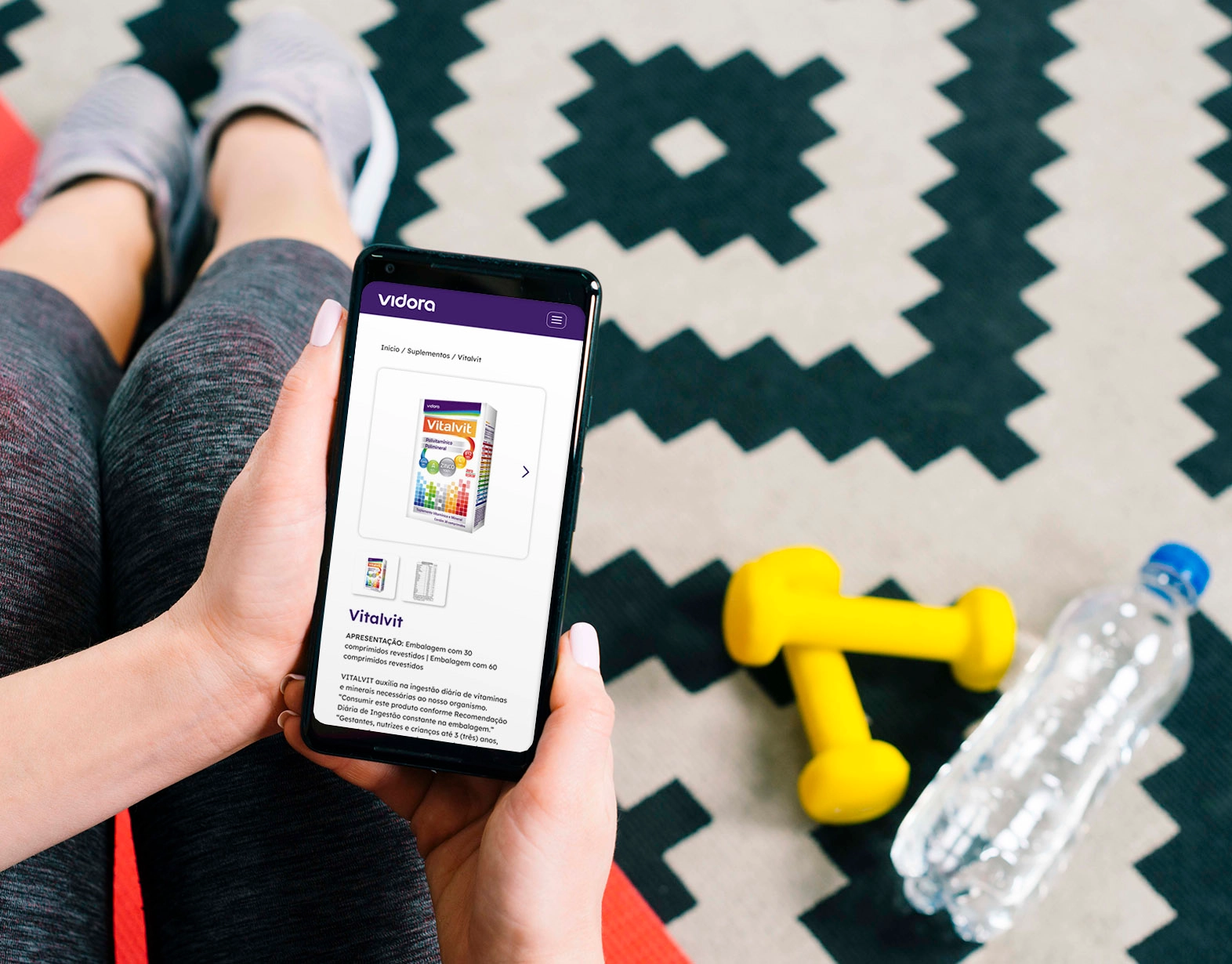 A person holding a smartphone displaying the product page for Vitalvit on the Vidora website. The page shows an image of the Vitalvit box, navigation breadcrumbs, and a detailed text description of the product. Next to the person's feet are a clear bottle of water and two bright
          yellow dumbbells.